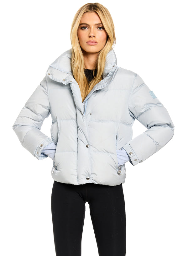 ANDI A blonde model wearing a light icy blue puffer jacket with a high collar and black pants, photographed against a solid white background.