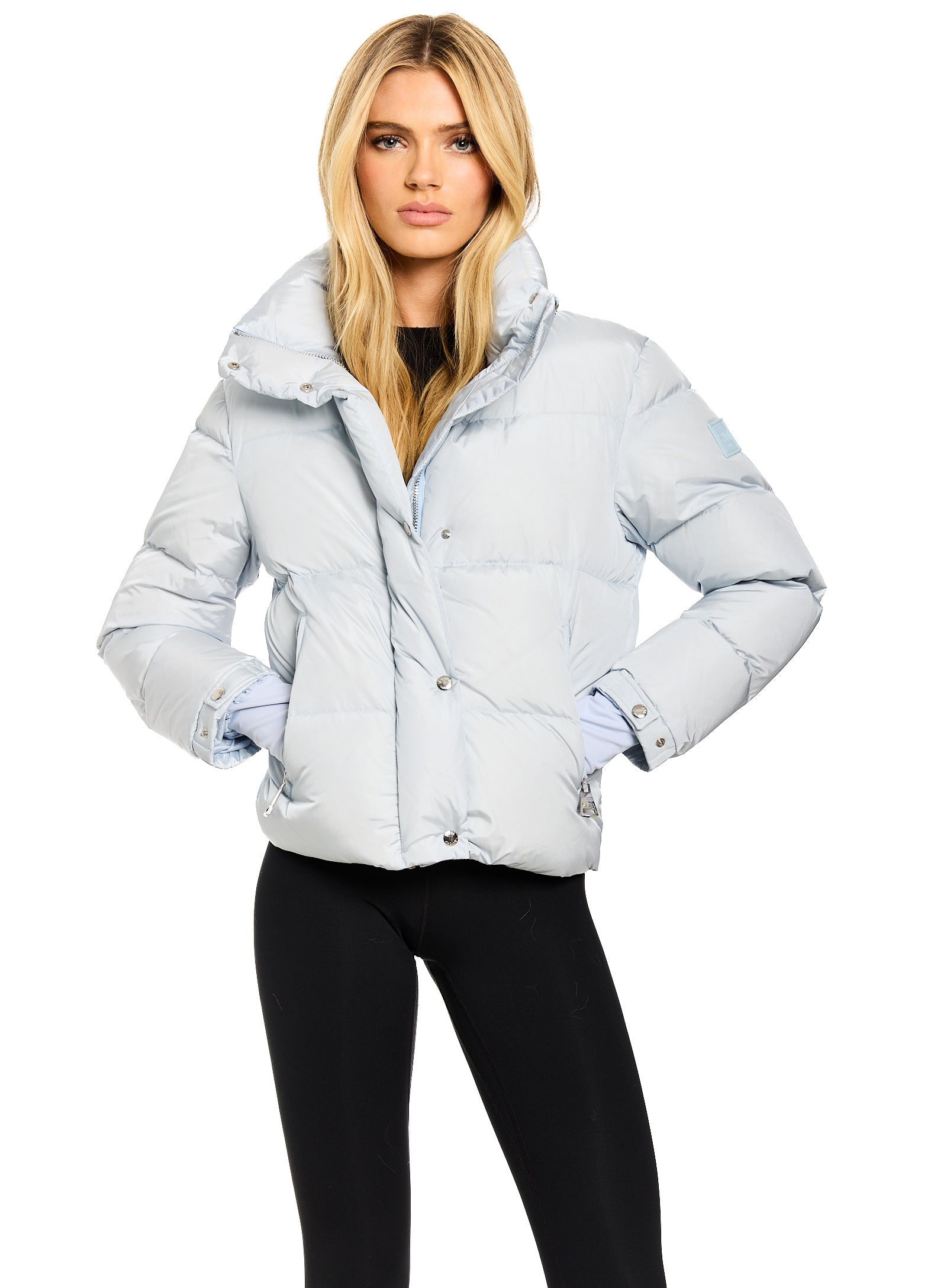 ANDI A blonde model wearing a light blue puffer jacket with a high collar and black leggings, standing against a solid white background.