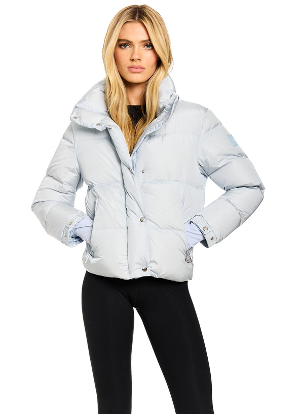 ANDI A blonde model wearing a light blue puffer jacket with a high collar and black leggings, standing against a solid white background.