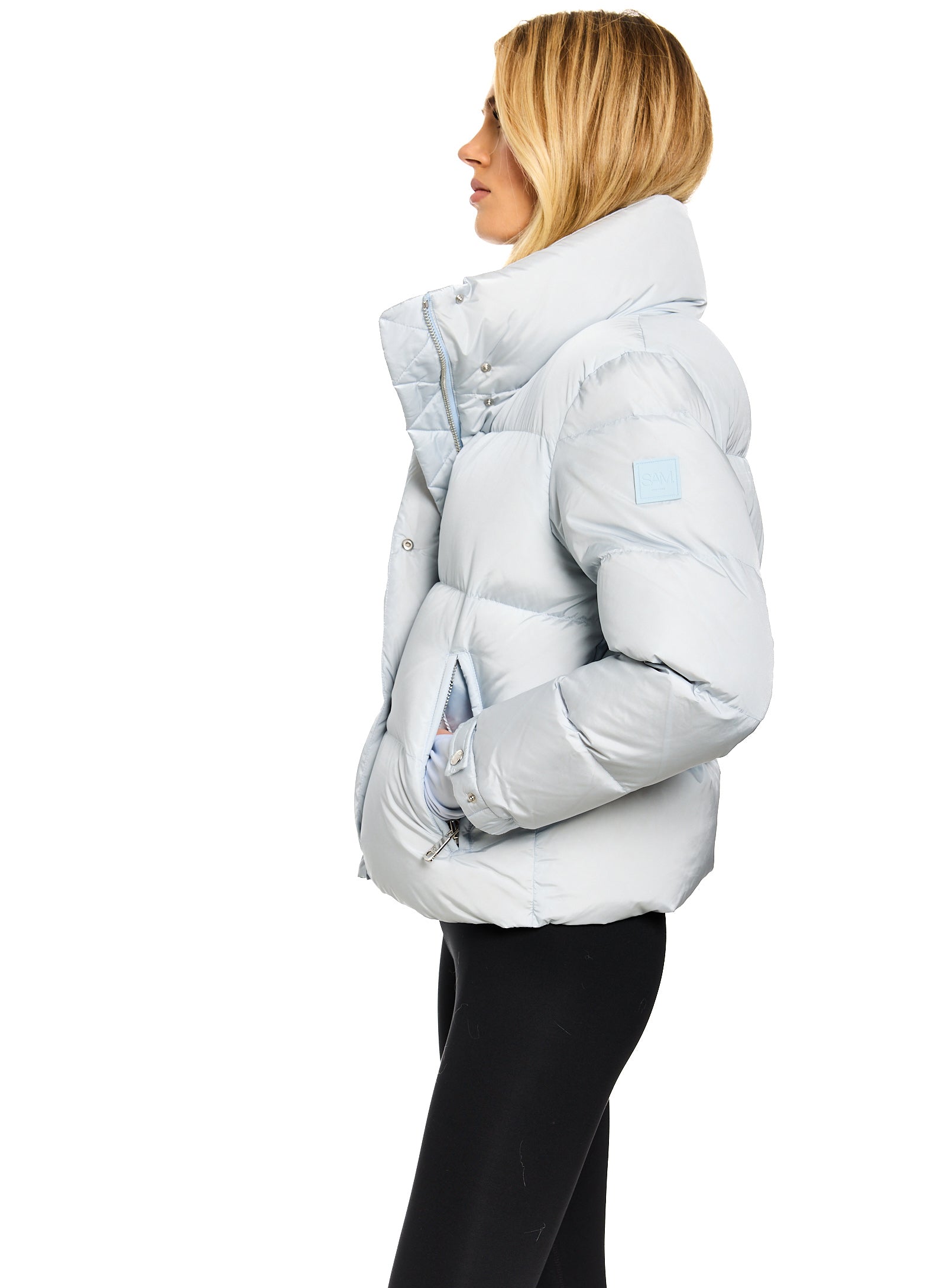 ANDI A woman shown in profile wearing a light icy blue puffer jacket with a high padded collar and black pants, photographed against a plain white background.