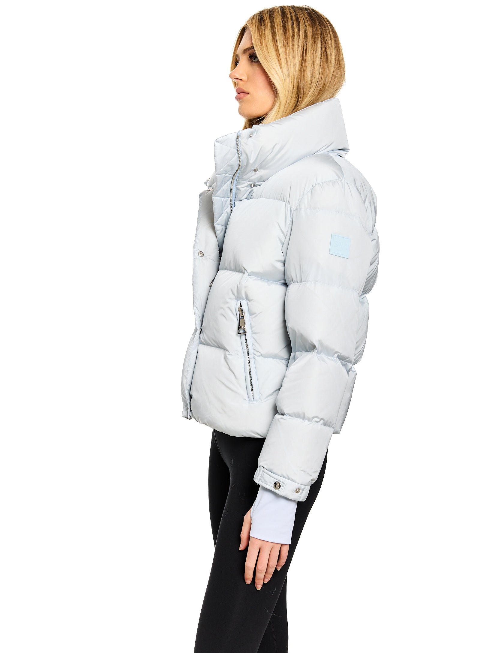 ANDI A woman shown in profile wearing a light icy blue puffer jacket with a high collar and black pants, photographed against a plain white studio background.