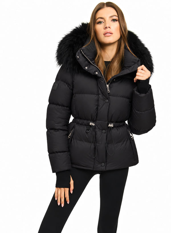 A woman modeling a black puffer jacket with a large black fur-trimmed hood, belted waist, and silver zipper details, set against a plain white background.