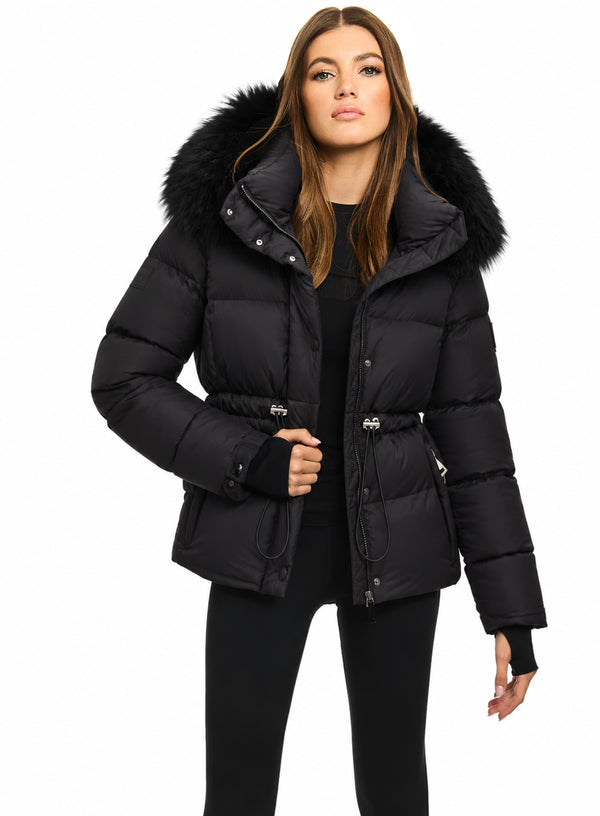 DELANEY A woman modeling a black puffer jacket with a large black fur-trimmed hood, belted waist, and front snap closure, photographed against a plain white studio background.